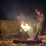 Experience the magic of Fraser Island as a man in a cowboy hat relaxes by a campfire on a Kgari Swag Adventures tour at night.