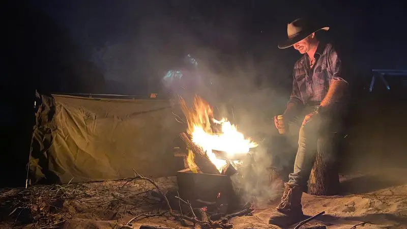 Experience the magic of Fraser Island as a man in a cowboy hat relaxes by a campfire on a Kgari Swag Adventures tour at night.