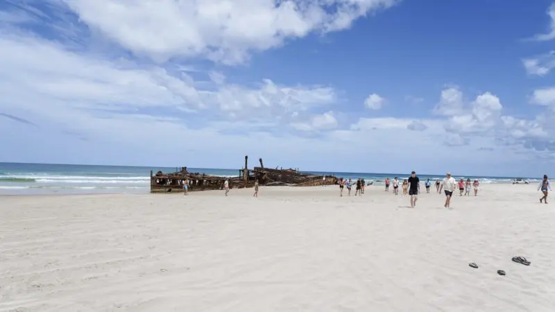 Tourists explore golden sands beside a historic rusted shipwreck on Fraser Island during the ultimate Kgari Safari 4WD camping experience.