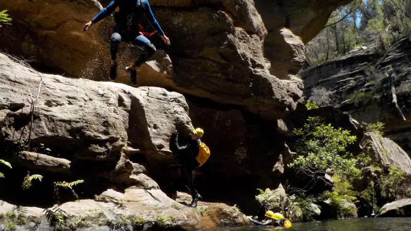 Three helmeted adventurers leap into a river on the Double Canyon Adventure, set in the scenic Blue Mountains wilderness.