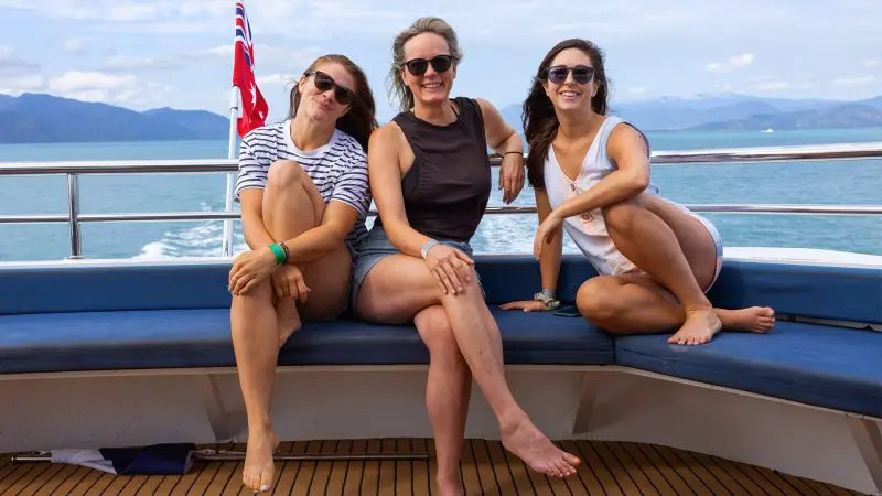 Three smiling women sit barefoot on a boat bench during their Evolution 2 Day Reef Cruise and Kuranda Train Skyrail adventure in Australia.