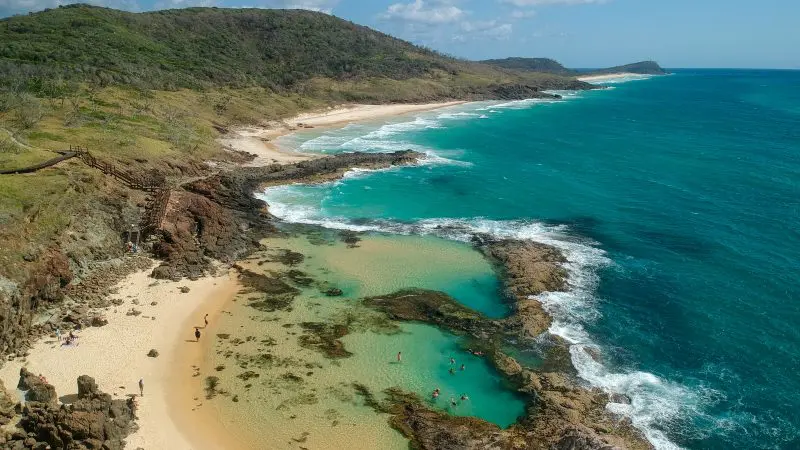 Stunning aerial view of Kgari 3 Day Getaway: rugged coastline, crystal-clear turquoise pools, golden sand beach, hills, rolling waves.