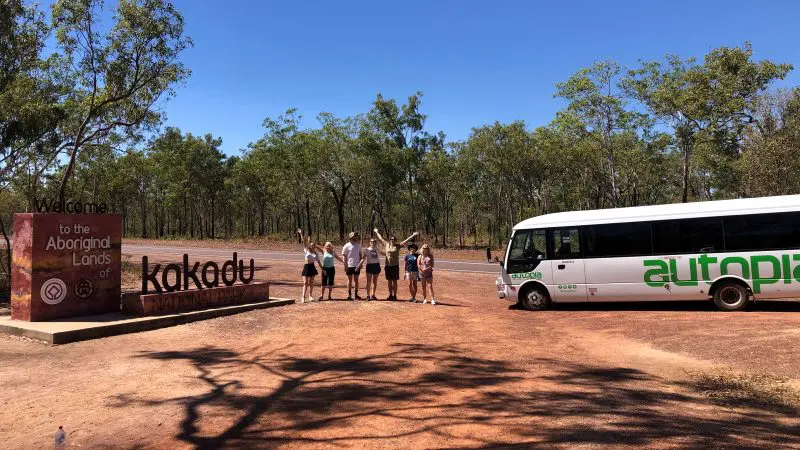 Visitors gather next to a “Welcome to Kakadu” sign and tour bus, set for a 1 Day Kakadu Wilderness Escape and Fogg Dam Wetlands tour.