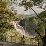 Person on platform overlooking powerful waterfall cascading into expansive wetlands amid lush, green hills; breathtaking scenic view.