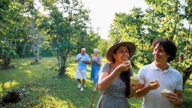 Two visitors enjoy ice cream and smile as an older couple strolls behind them in a sunlit Cape Tribulation Daintree orchard.