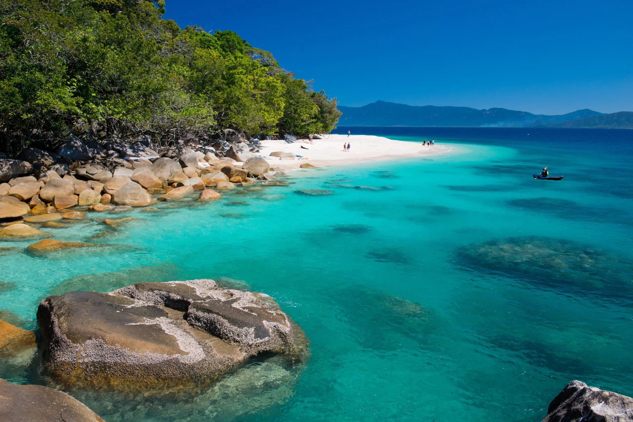 Pristine white sandy beach on Fitzroy Island, turquoise water, rocky shoreline, and lush trees beneath a crystal-clear sky.