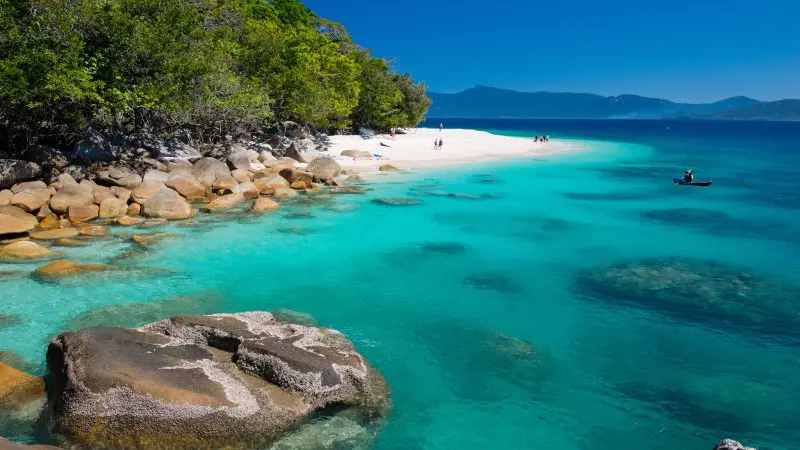 Pristine white sandy beach on Fitzroy Island, turquoise water, rocky shoreline, and lush trees beneath a crystal-clear sky.