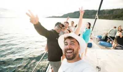 Happy friends capture a selfie on a Kiwi Classic sailboat, flashing peace signs with scenic blue waters and lush green hills behind them.