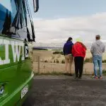 A diverse group of people stands on a paved road beside a vibrant green coach, ready for travel or tours, under clear daylight.
