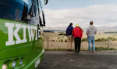 A diverse group of people stands on a paved road beside a vibrant green coach, ready for travel or tours, under clear daylight.
