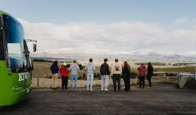 A group of hikers stand on a scenic carriageway, gazing at the breathtaking Kiwi Classic mountains under a clear New Zealand sky.