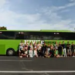Diverse group posing in front of a vibrant green bus, performing the Funky Chicken pose for a fun, memorable photo opportunity.