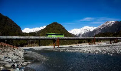 A city bus crosses a scenic bridge spanning a wide river, with urban skyline and lush greenery visible in the background.