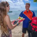Smiling woman in snorkelling gear interacts with uniformed man holding Fitzroy Island Essentials life jacket on sunny beach.
