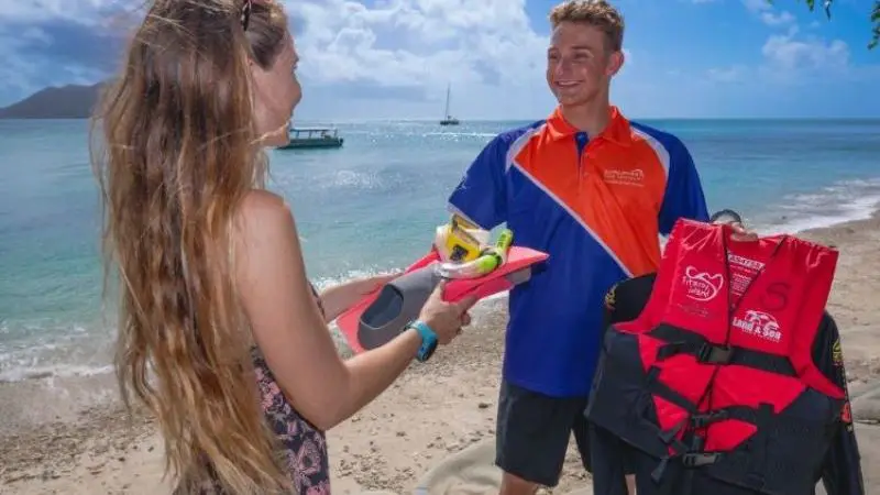 Smiling woman in snorkelling gear interacts with uniformed man holding Fitzroy Island Essentials life jacket on sunny beach.