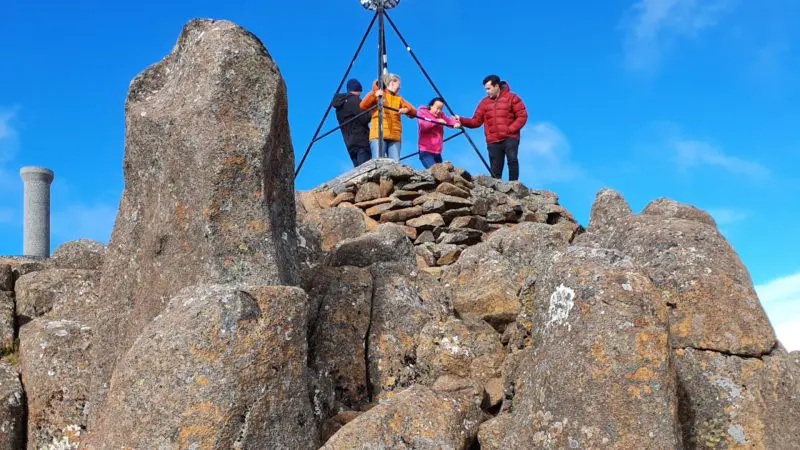 Four hikers atop Mount Wellington’s rocky summit during a guided morning walking tour, framed by a vivid blue sky and scenic views.