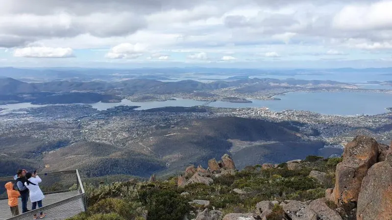 Visitors savour panoramic views during a Mt Wellington Afternoon Driving Tour, overlooking Hobart city, Derwent River, and rolling hills below dramatic clouds.