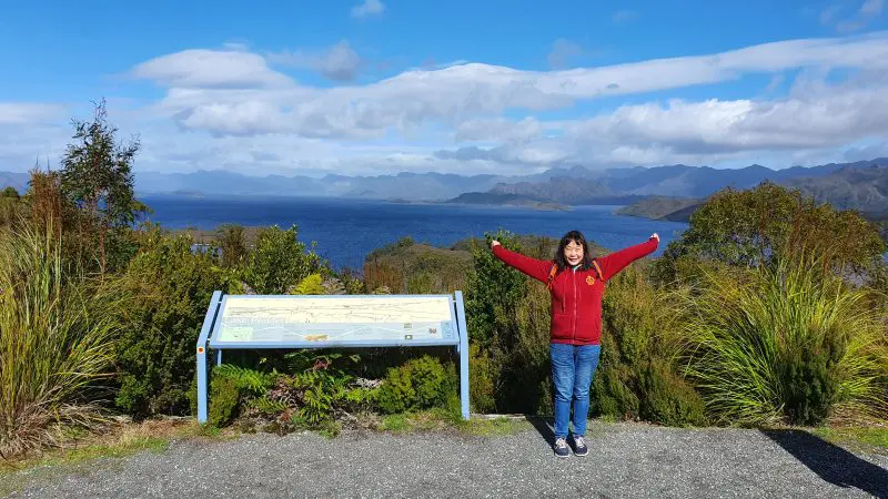 Woman in vibrant red jacket joyfully raises arms beside scenic Lake Pedder during Gordon Dam Wilderness Small Group Day Tour Tasmania.