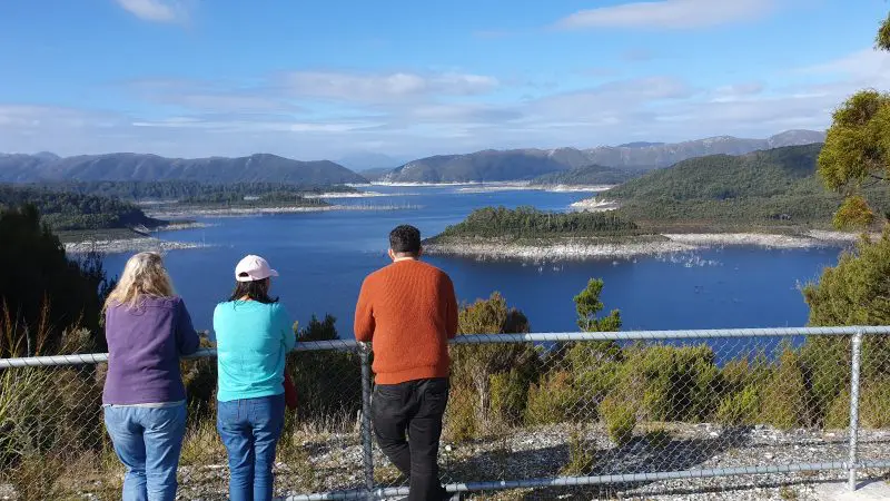 Three travellers by a metal fence gaze at scenic Lake Pedder on a guided Small Group Day Tour, surrounded by pristine Tasmanian wilderness.