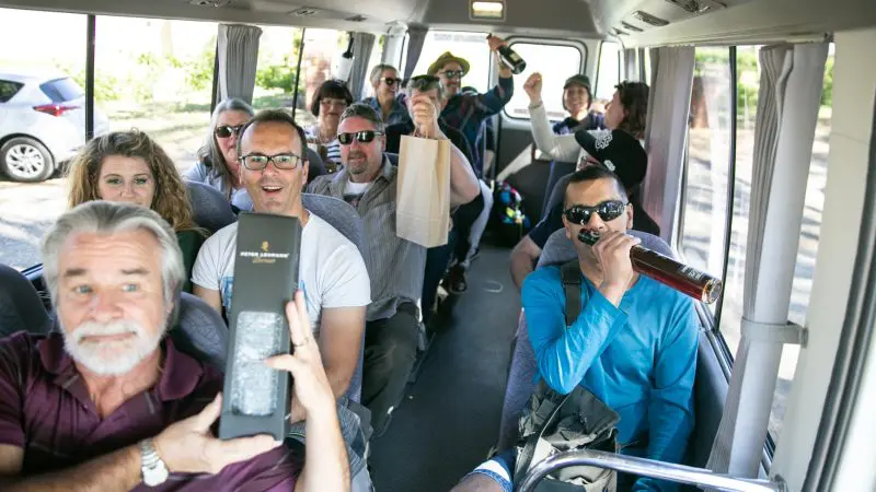 Smiling friends enjoy drinks and gifts on a Taste The Barossa Premium Wine Tour coach, celebrating with local wines and scenic views.