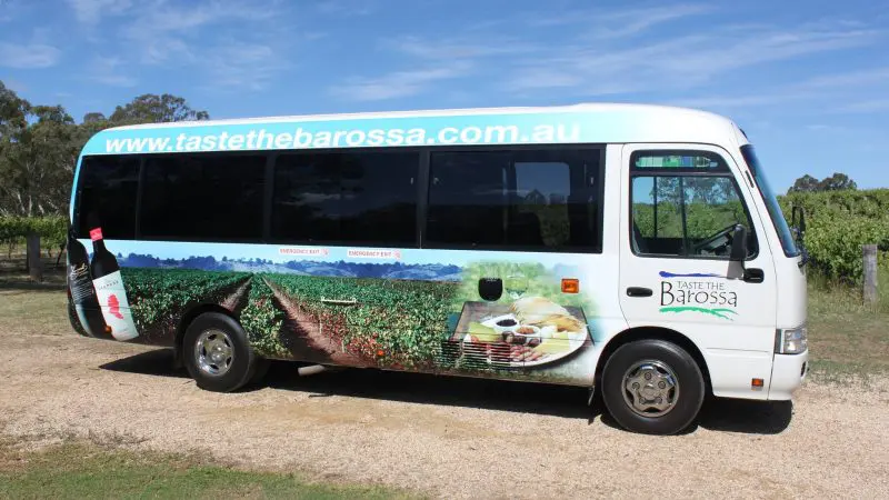 Taste The Barossa wine tour bus with vibrant vineyard graphics on a rural dirt track, advertising premium Barossa Valley tours.