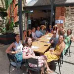 Friends savour wine and cocktails at a bright outdoor café, unwinding post 1 Day Wine Tasting Coast Afternoon Tour by the seaside.