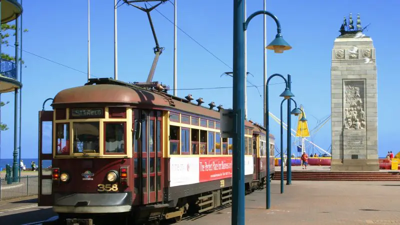 Vintage red tram passing a historic stone monument by the coastal road on a sunny 1–2 day wine tasting tour afternoon, scenic view.