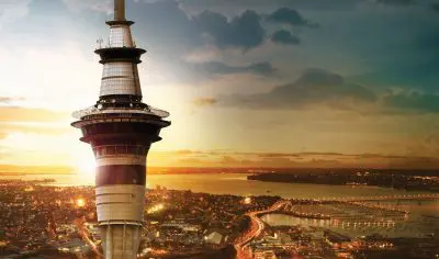 Sky Tower rising above Auckland City at sunset, vibrant lights illuminating streets and waterfront, stunning panoramic cityscape view.