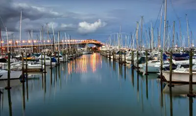 Sailboats berthed at Auckland marina at dusk, with city skyline, illuminated bridge, and vibrant lights reflecting on tranquil water.