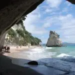 Breathtaking Coromandel coastal scene: golden sandy beach and turquoise sea framed by a rocky cave, people exploring the shore.