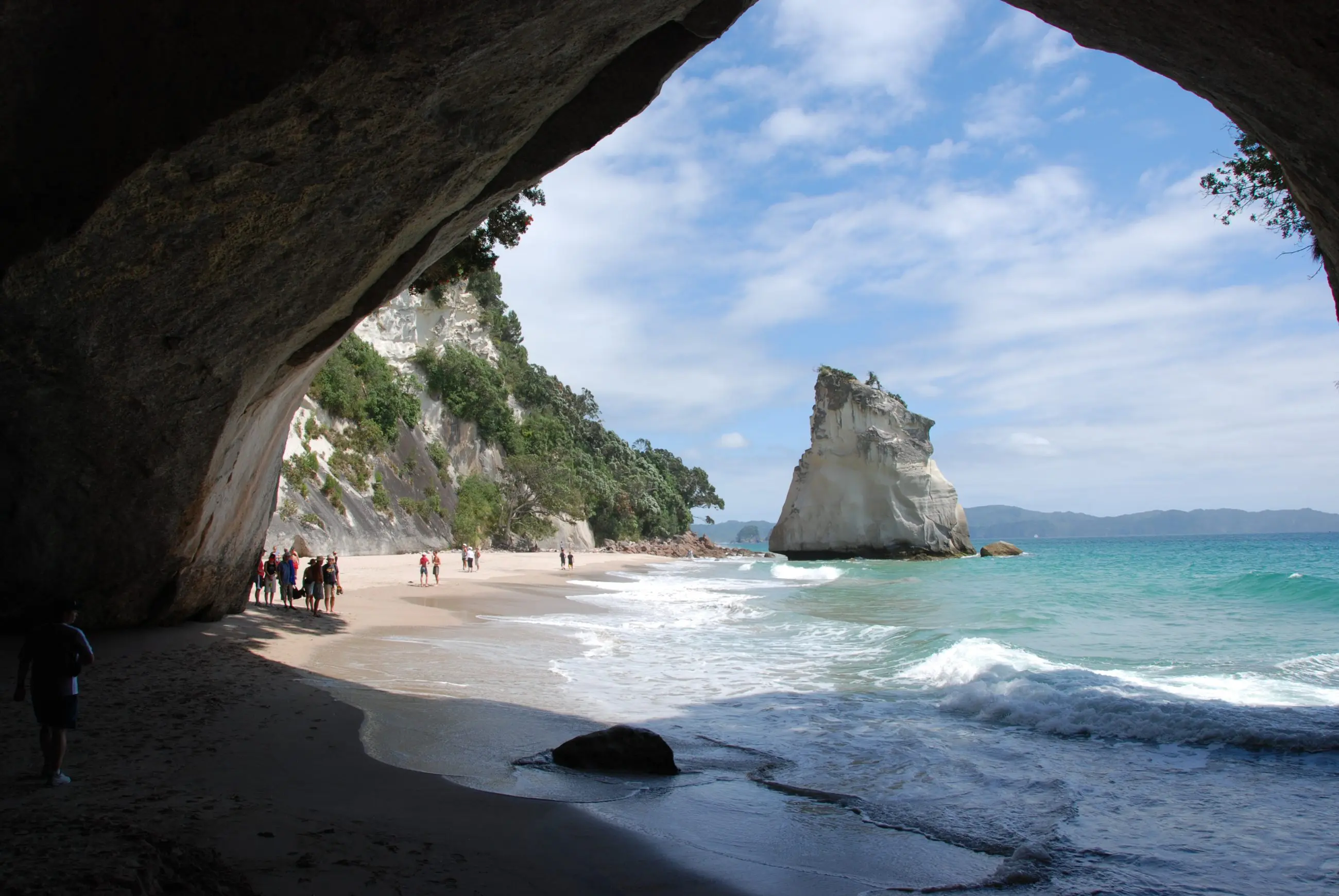 Breathtaking Coromandel coastal scene: golden sandy beach and turquoise sea framed by a rocky cave, people exploring the shore.