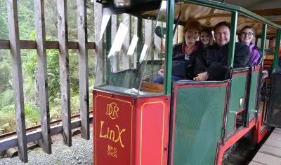 Happy group of four riding a scenic green and red railway carriage at an outdoor Coromandel station, enjoying a vibrant New Zealand tour.