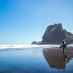 Surfer with board walks along the wet, scenic West Coast beach near crashing waves and a rocky hill beneath clear blue sky.