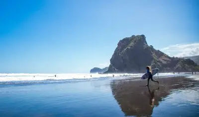Surfer with board walks along the wet, scenic West Coast beach near crashing waves and a rocky hill beneath clear blue sky.
