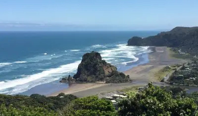 Breathtaking West Coast beach with rocky outcrop, rolling waves, lush greenery, and scenic hills beneath a vibrant blue sky.