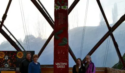 Four visitors stand beside a vibrant red totem pole inside the glass-domed Waitomo Caves Centre, surrounded by wooden floors and exhibits.