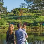 A family enjoys the scenic Hobbiton Movie Set with iconic hillside hobbit houses by a tranquil pond under a clear blue sky in New Zealand.