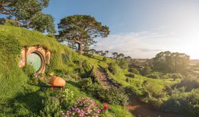 Picturesque round green door at Hobbiton Movie Set on a lush grassy hillside, vibrant flowers, trees, and stone path at sunrise.