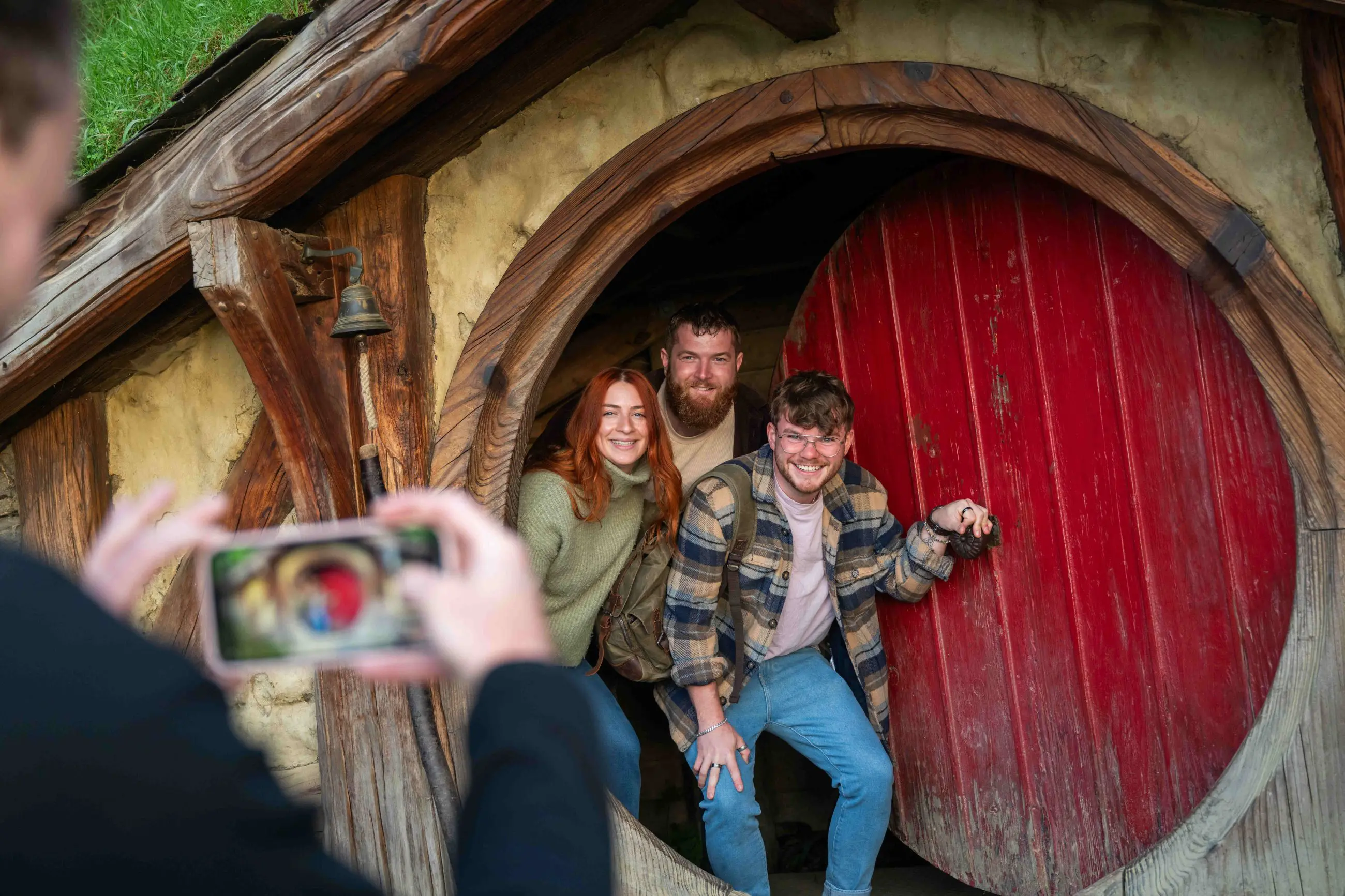 Three happy visitors pose by a red Hobbit door at the Hobbiton Movie Set as their photo is captured on a smartphone for memories.