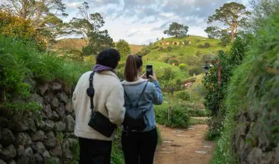 Visitors walk along a scenic path at Hobbiton Film Set, capturing the iconic lush green hillside and distinctive round hobbit doors nearby.