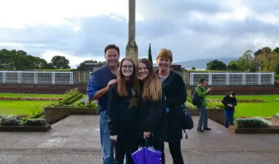 Family of four on a private Auckland tour, smiling by a tall monument with a vibrant purple umbrella under overcast skies.