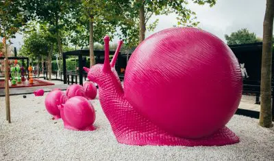Striking large pink snail sculptures and smaller snails adorn Matakana park, enhancing scenic white gravel paths amid lush green trees.