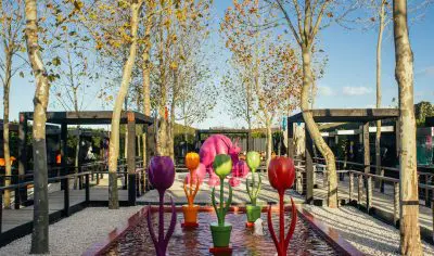 Vibrant tulip sculptures by a tranquil Matakana Art waterway, surrounded by lush trees, walkways, and a striking pink Wine Village sculpture.