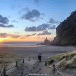 Individual strolling along a sandy footpath toward Piha Beach at sunset, dramatic cliffs and vibrant clouds in the scenic background.