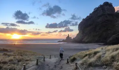 Individual strolling along a sandy footpath toward Piha Beach at sunset, dramatic cliffs and vibrant clouds in the scenic background.