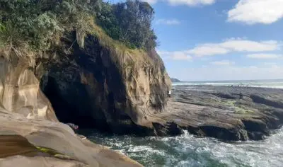 Muriwai Beach rocky shoreline with dramatic sea cave, lush green cliffs, rolling ocean waves, and a pristine sky ideal for private tours.