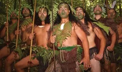 Six Māori warriors with intricate tattoos and spears stand in a lush forest, set to welcome guests to an immersive Māori Dinner Show.