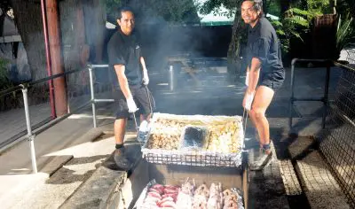 Two people lower trays of steaming meats and vegetables into a traditional Māori earth oven outdoors during an authentic evening feast.