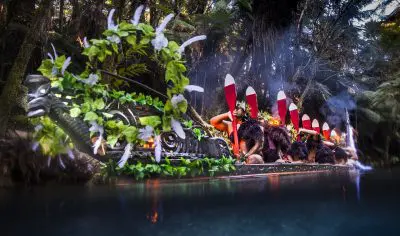 Experience authentic Māori culture as performers in traditional attire paddle an ornately carved waka at dusk during a magical evening performance.