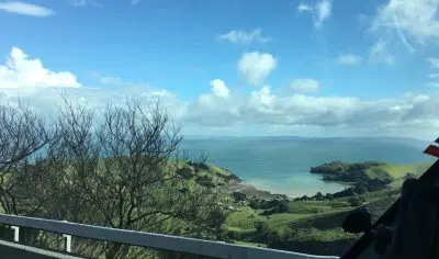 Scenic view of Coromandel Beach from roadside featuring lush green hills, turquoise blue sea, and clear sky under bright sunlight.